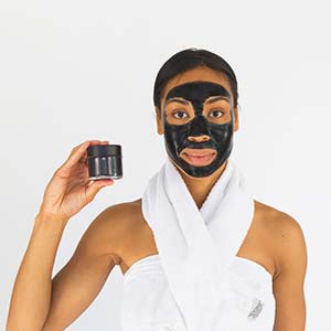 Photo of a woman with a charcoal mask on her face and holding a bottle of the product. Click on the image for more information about DIY charcoal mask. 